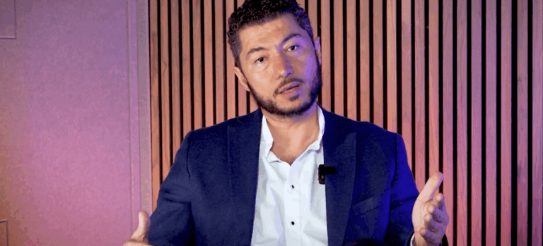 Man in a navy blazer and white shirt speaks, gesturing with his hands in a studio with vertical wooden slats behind him.