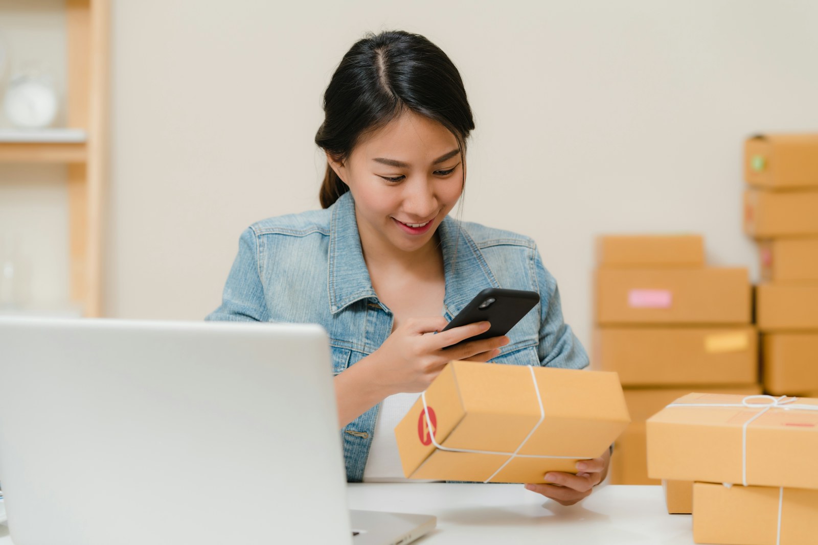Ecommerce seller checking an order on a phone beside a laptop and packed shipping boxes