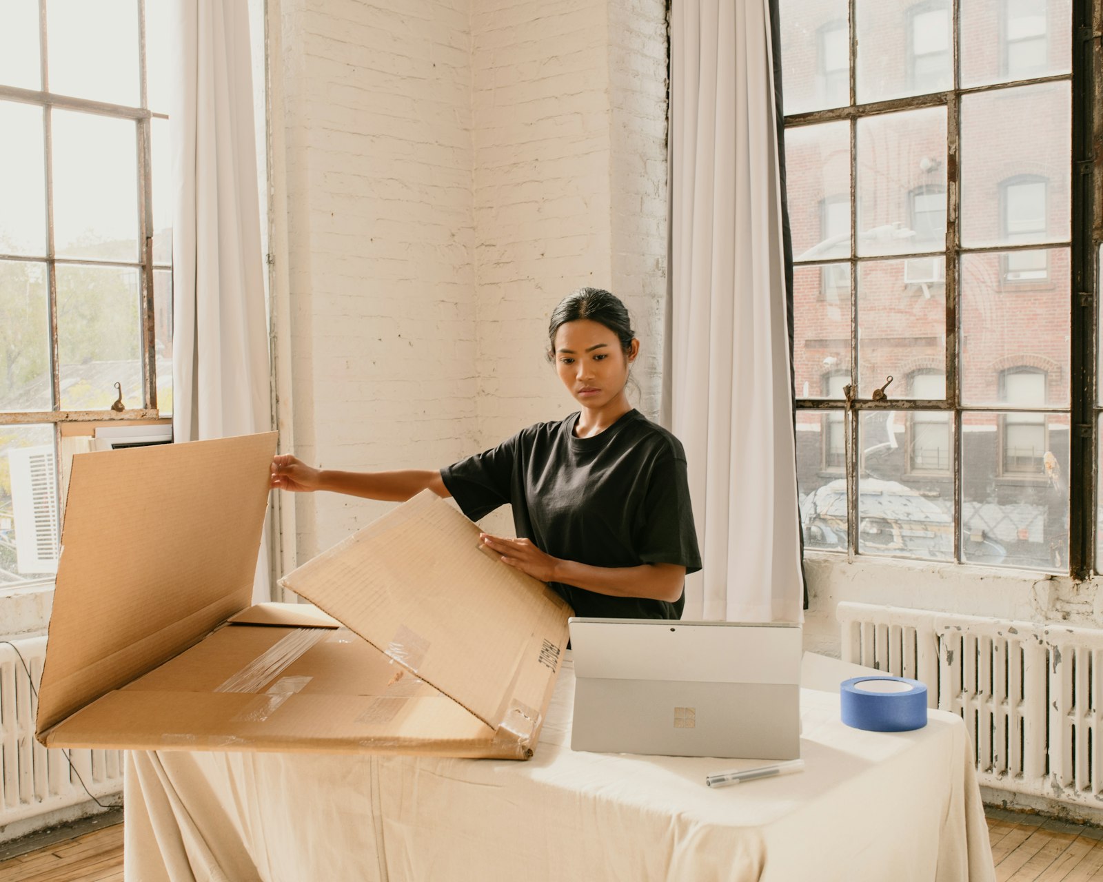 Store owner opening a cardboard shipping box next to a laptop by a large window